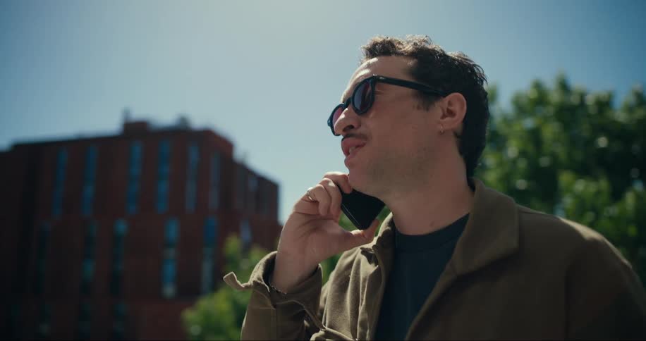A man wearing sunglasses talks on a smartphone under a clear blue sky on a sunny day