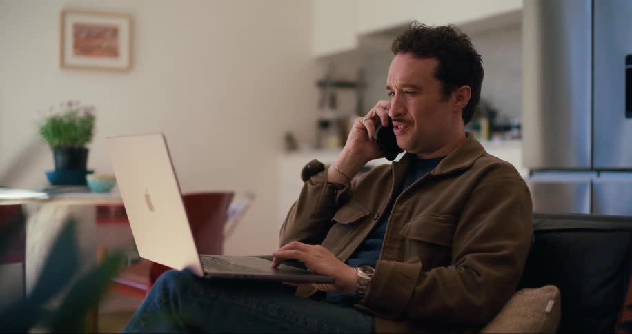 A man is sitting on a sofa using a laptop then picks up a phone and starts talking to it in his ear