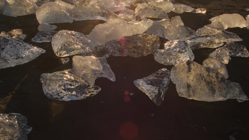 Scattered pieces of translucent ice glisten on a dark surface reflecting light and revealing a faint watermark