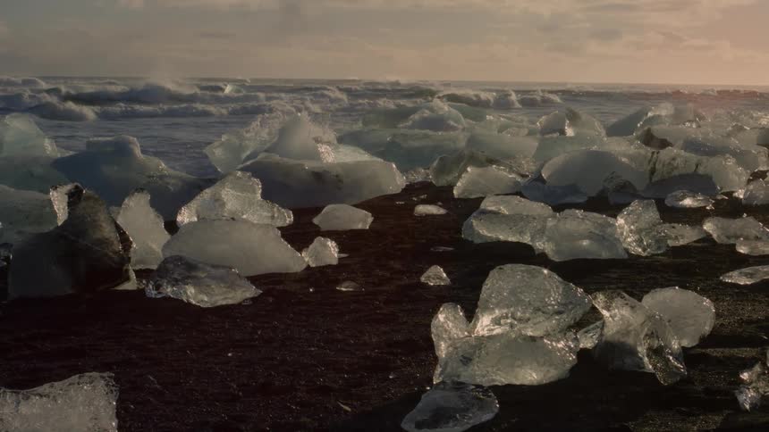 Large translucent chunks of ice rest on a black sand beach with the ocean churning in the background under a cloudy sky