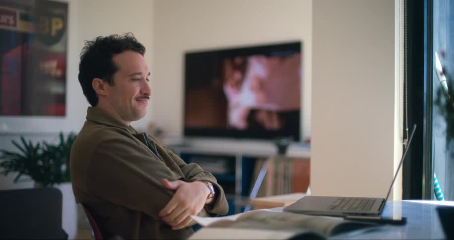 A man in a casual brown jacket is sitting indoors leaning back with his hands behind his head smiling slightly with a laptop in front of him and a television in the background