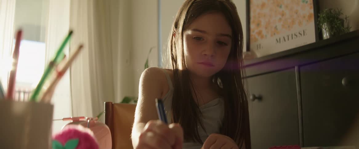 A girl with long hair is sitting at a table drawing with colored pencils in a sunlit room with a window