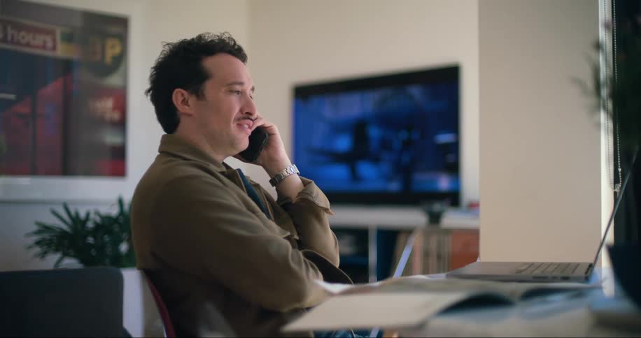 A man in a casual brown jacket is sitting indoors holding a phone to his ear while speaking with a laptop in front of him and a television in the background