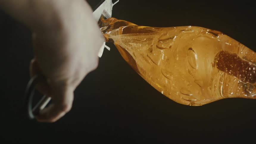 A hand holds a tool shaping a molten piece of ambercolored glass against a dark background