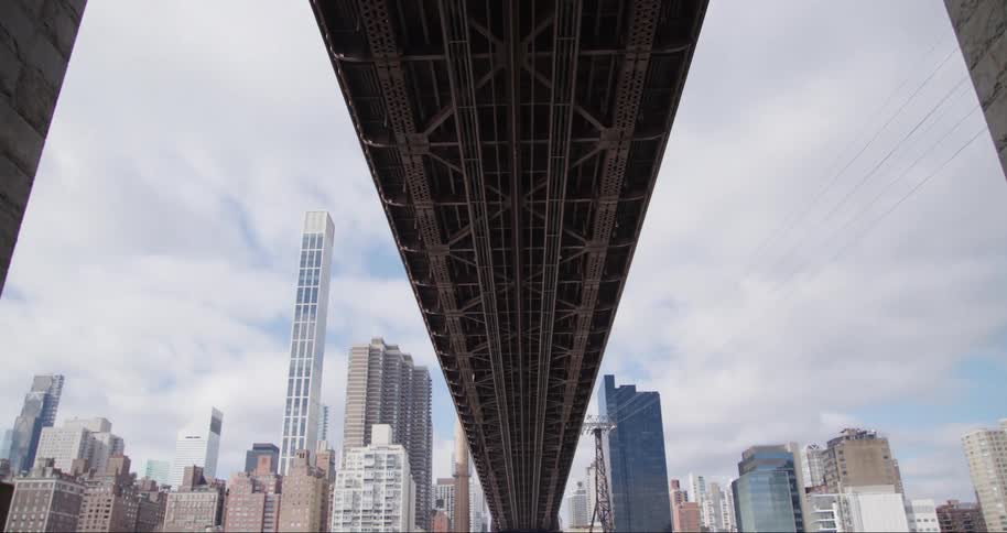 A low angle shot from below the queensboro bridge captures its intricate steel structure extending over the east river with the new york city skyline and the distinctive 59th street bridge ed koch queensboro bridge in the background