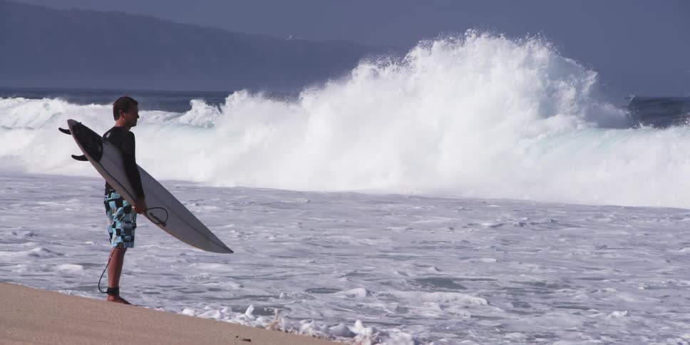 A surfer stands on a sandy beach and watches the waves