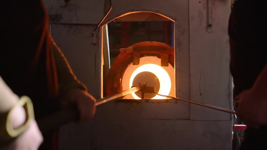 Two individuals are using long metal tools to manipulate molten glass inside a glowing hightemperature furnace