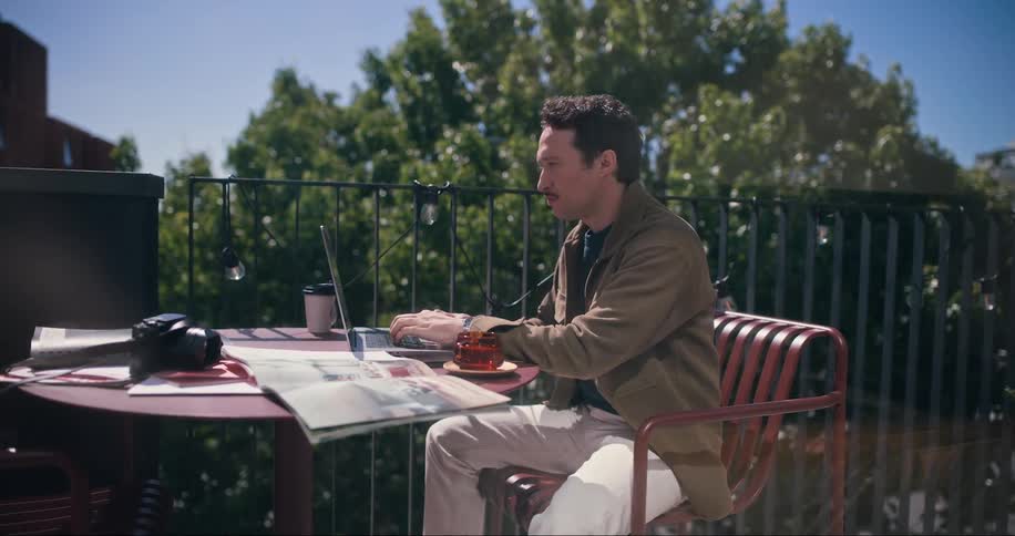 A man typing on a laptop at a table on a sunny balcony