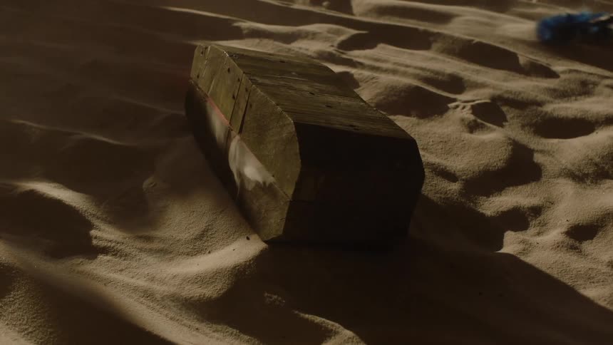 A dark rectangular block with a subtle red line lies on a sandy surface casting a long shadow