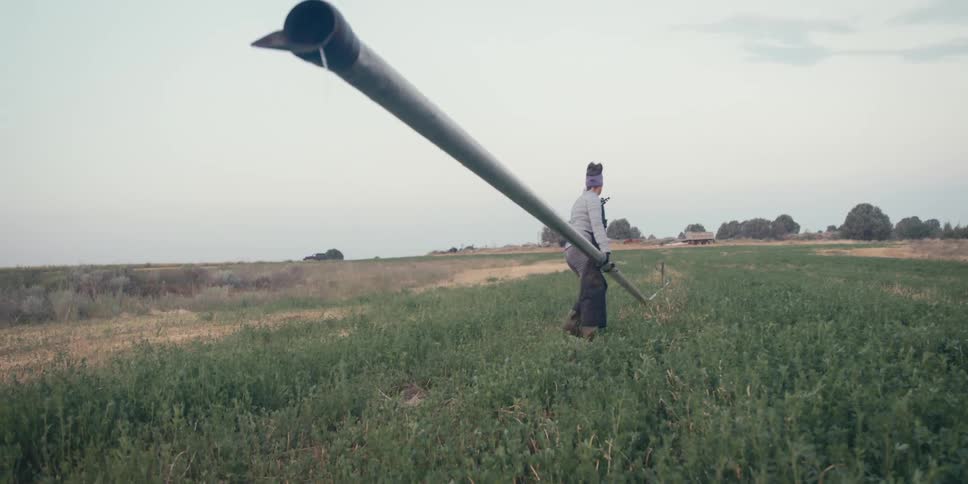 A farmer adjusts an irrigation pipe in a wide alfalfa field at sunset surrounded by open farmland and distant trees under a soft evening sky