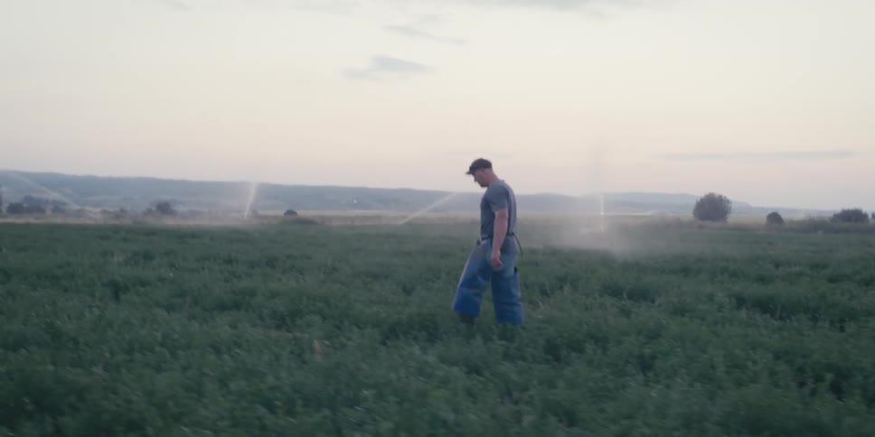 A man in a cap walking in a field with some sprinklers spraying water to the sides in the background