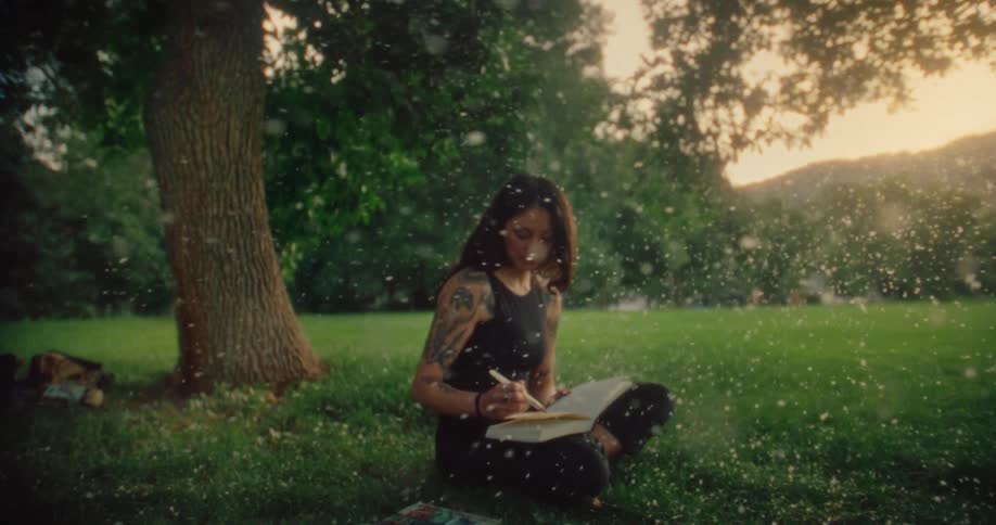 A woman with long hair and tattoos on the left arm is writing on a notebook outdoors with sunlight filtering through the trees and white particles floating around her