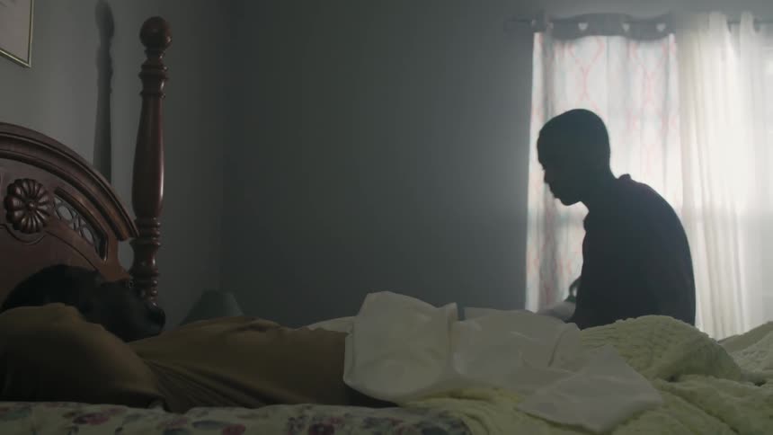 A boy sits down next to a man laying down on a bed