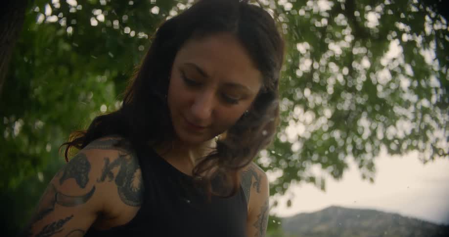A woman with long hair and tattoos on the left arm is writing on a notebook outdoors with sunlight filtering through the trees and white particles floating around her