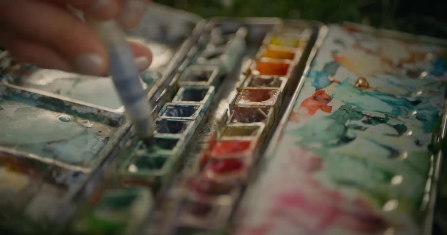 A hand holding a water brush mixes colors in a wellused watercolor palette filled with an array of hues