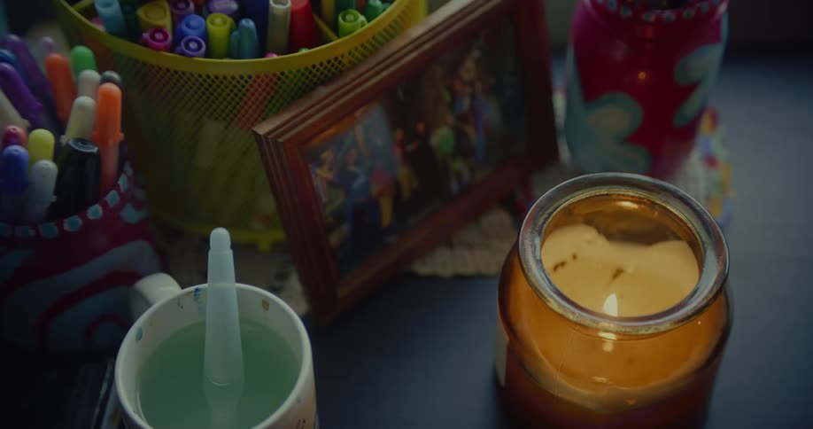 A lit candle in an amber glass jar sits among colorful markers pens and a framed picture on a dark surface
