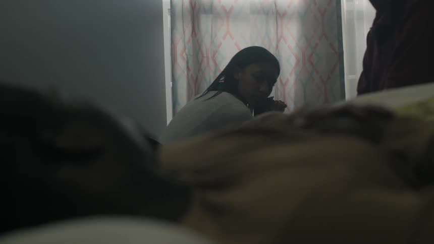 Man lying on his back in bed talking with another person that sits close to a window