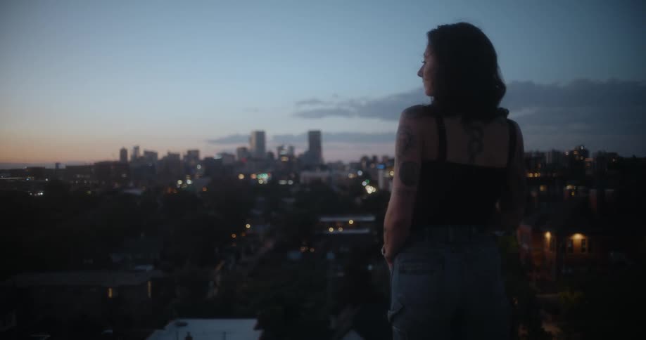A woman with tattooed arms and back stands outside gazing at the city skyline at sunset