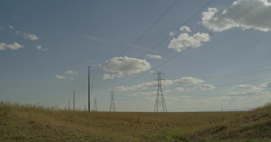 A vast dry field stretches under a blue sky dotted with fluffy white clouds bisected by tall electrical pylons and their connecting wires