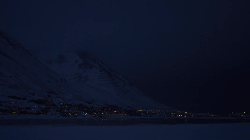 House lights shine in a town located between the water and a snowy mountain on a winter night