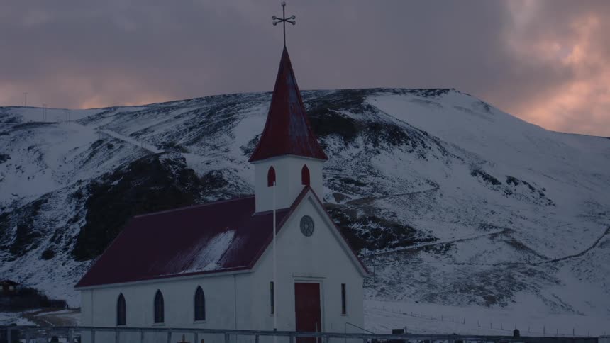 A white red roofed chapel in front of a snow covered hillside under a clouded sky at dusk