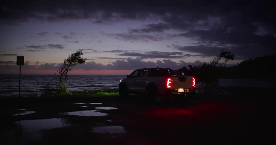 A white pickup truck is parked near a beach at dusk and two people climb into the back