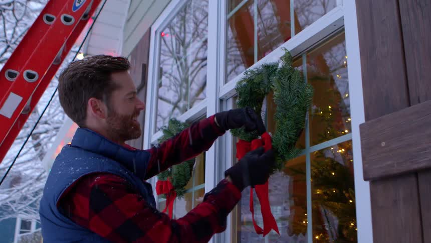 Man hangs a christmas wreath on a window during a snowfall