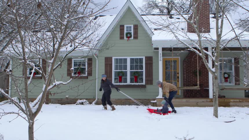 A mother and father take their toddler son sledding in their front yard at christmastime