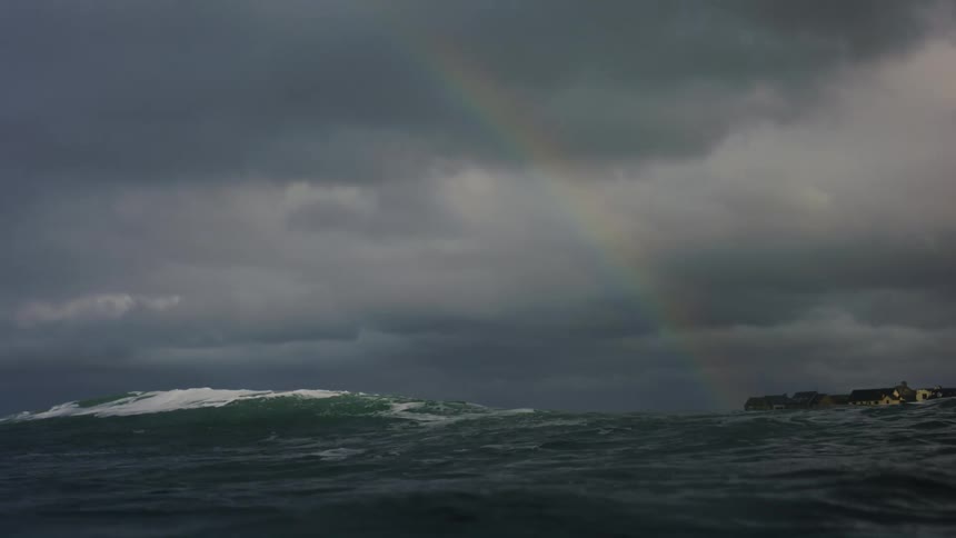 A faint rainbow arches over a dark choppy sea beneath a heavy overcast sky with a coastal town on the horizon
