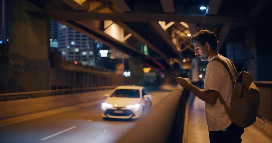 Man standing on the sidewalk under the bridge uses his phone while traffic passes by