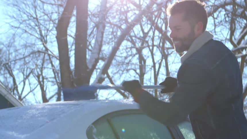 A man brushes ice from the roof of a car parked in a neighborhood on a sunny winter morning