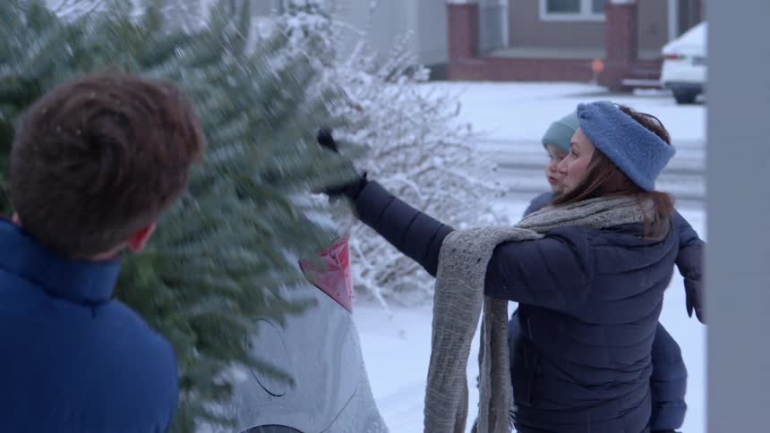 A man unloads a christmas tree from the roof of a snowcovered car while his wife holding their son assists him with houses and holiday lights in the background