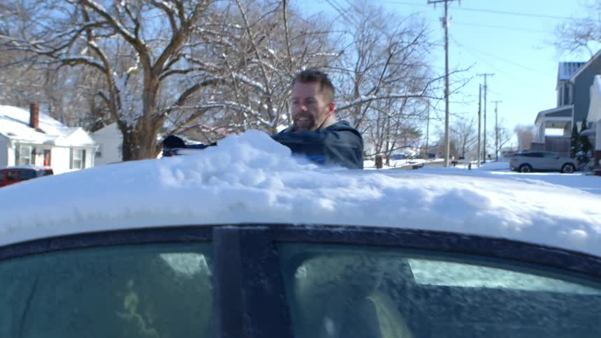 A man brushes the snow from the roof of a car parked in a neighborhood on a sunny winter morning