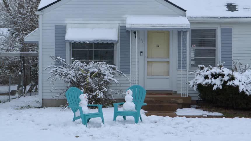 Two small snowmen sit in turquoise chairs in front of a snow covered house