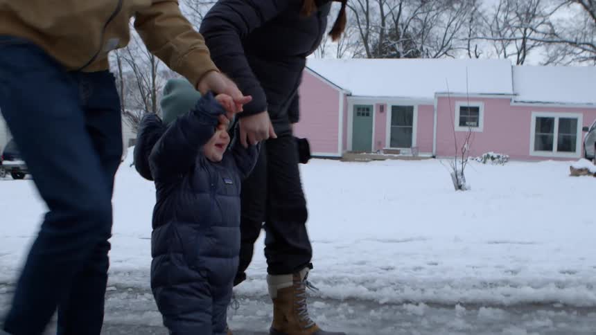 Two adults and a child all dressed in winter clothing walk handinhand on a snowcovered street in front of a pink house