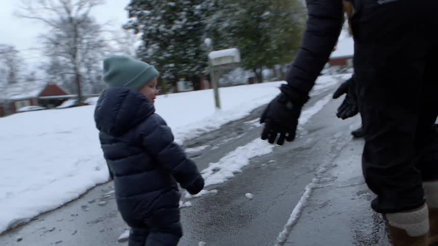 Some adults accompanying a baby who is walking on a street that is covered in ice and snow and everyone is wearing warm clothes