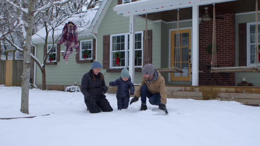 Parents with their young son in the backyard of a snowy house preparing to make a snowman