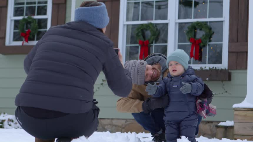 Dad and baby posing on the snow in front of house while mom takes a picture