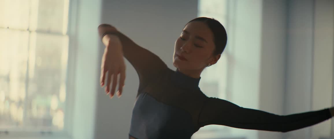 A woman dances ballet in a welllit room with large windows wearing a dark longsleeved outfit with an open back