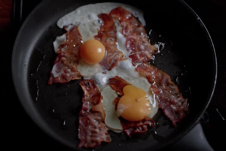 Pair of hands putting an egg to fry in a frying pan with bacon