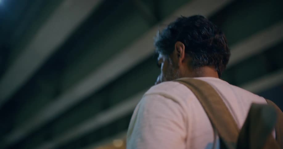 A person with short curly hair is seen from behind wearing a white shirt and carrying a backpack walking under a bridge at night