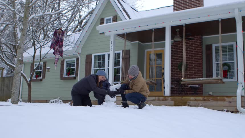 A family builds a small snowman in front of a house with a green exterior and a porch