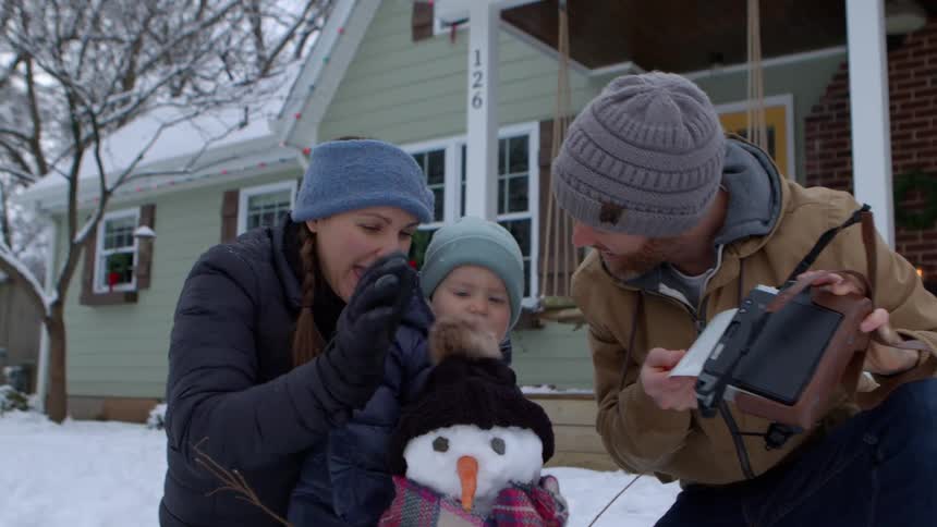 Dad taking a selfie with a polaroid camera with mom and baby behind a snowman