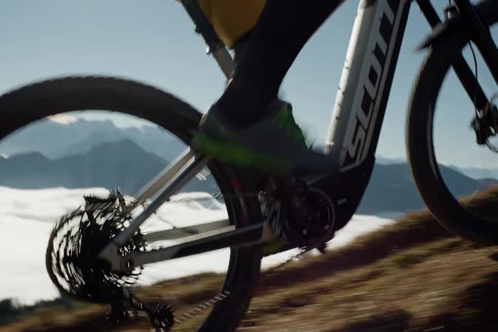 A closeup of bicycle wheels climbing a pathway over a mountain landscape partially obscured by clouds