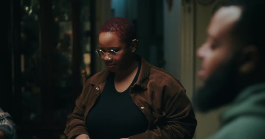 A woman with short hair and wearing glasses is looking down where there is a table and in front of it some people talking in an indoor environment