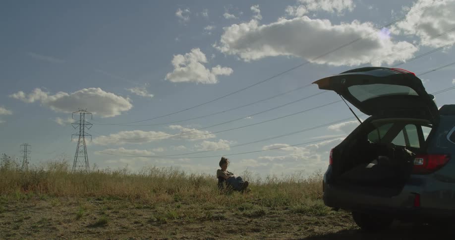 A woman sits in a folding lawn chair in a grassy field with a gentle breeze her car is parked nearby with the hatch open on a sunny day