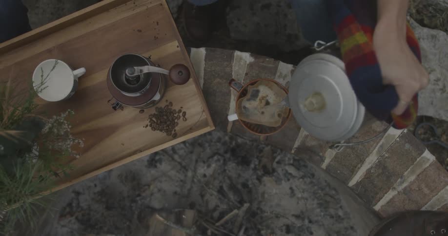 A person is pouring from a kettle near a campfire