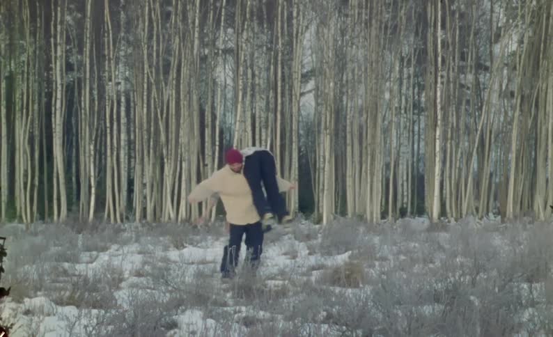 A person wearing a beige jacket and red beanie is carrying another person over their shoulder while spinning around in a snowy field with a dense forest of tall leafless trees in the background