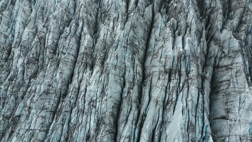 A textured glacier surface stretches out revealing a pattern of cracks and crevices in shades of gray and white