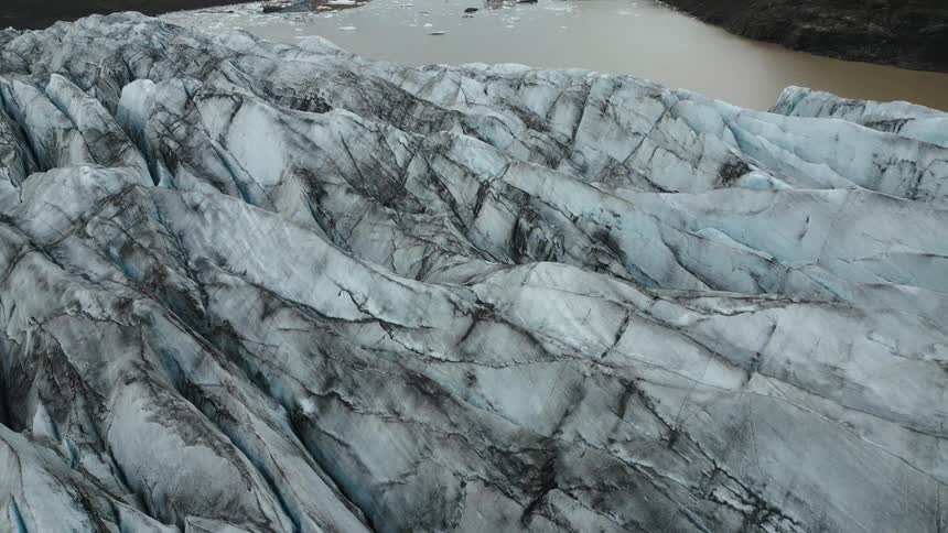 A glacier with prominent dark streaks and crevices leading to a muddy glacial lake surrounded by rugged terrain and distant mountains
